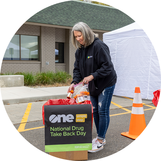 Woman with silver hair in black sweatshirt is placing pills into take back day box_circle frame.
