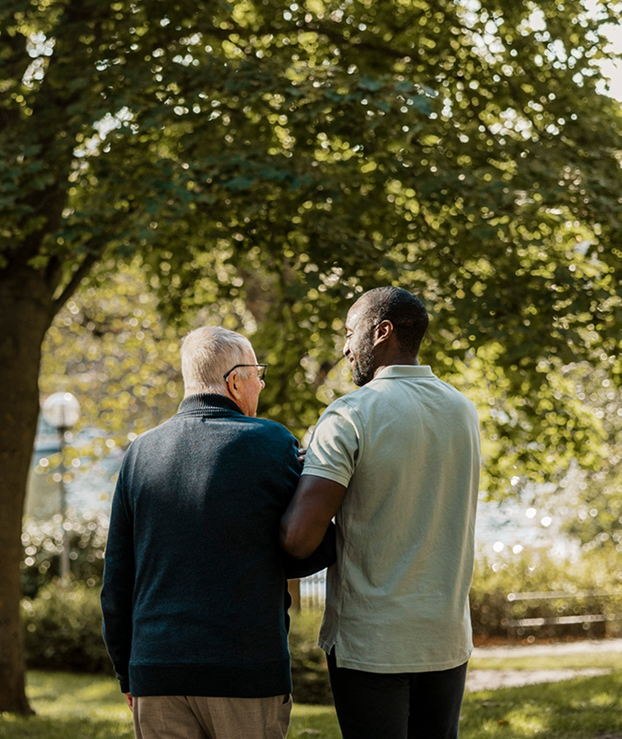 Younger man in green polo shirts is helping an older man in a grey turtle neck walk through a park.
