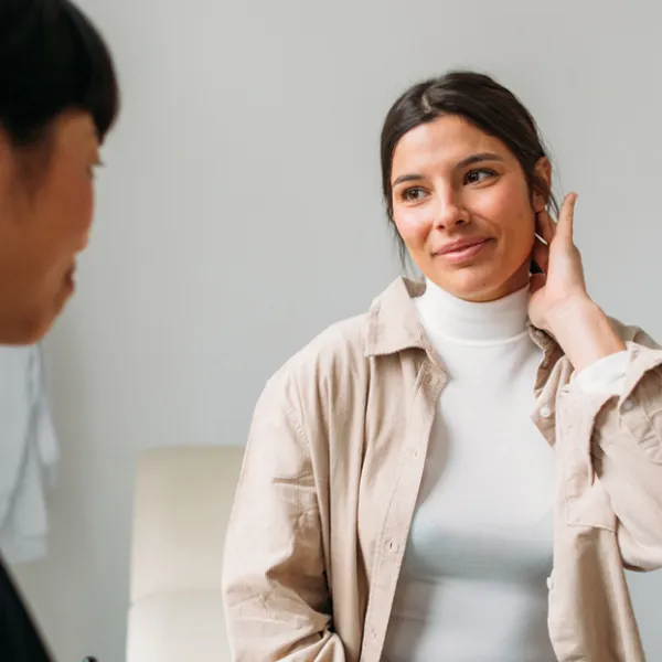 physician with black hair looks at female patient in white turtle neck with tan jacket.