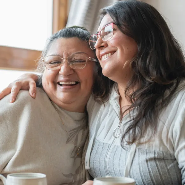 two women in glasses are hugging and smiling with coffee cups on the table in front of them.