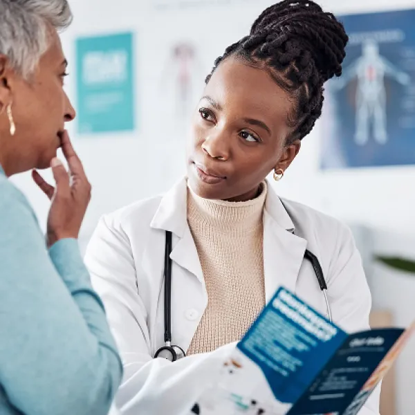 Doctor with a braided bun shows a rack card to a woman in a turquiose blue sweater with her hand up to her face.
