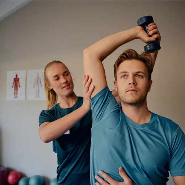 Man in blue shirt is holding dumbell over his head while being guided by a phsycian with a blonde pony tail.