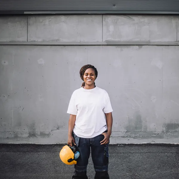 Person in white t-shirt with a braided pony tail is smiling at the camera holding a yellow hard hat in front of a concrete wall