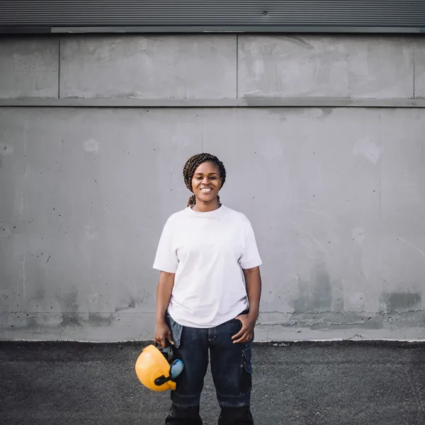 person in white t-shirt with a braided pony tail is smiling at the camera holding a yellow hard hat in front of a concrete wall.